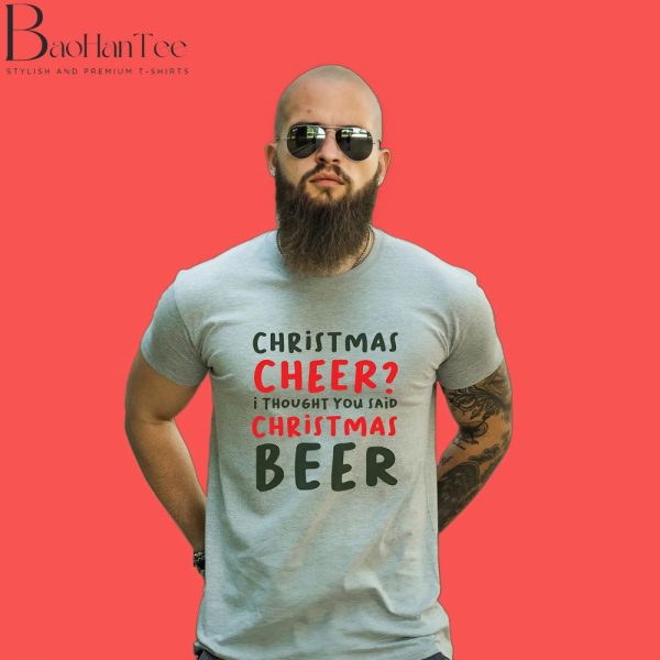 A man wearing a gray T-shirt with a festive slogan that reads "Christmas Cheer? I thought you said Christmas Beer" in black and red text. He has a full beard, sunglasses, and stands against a vibrant red background.