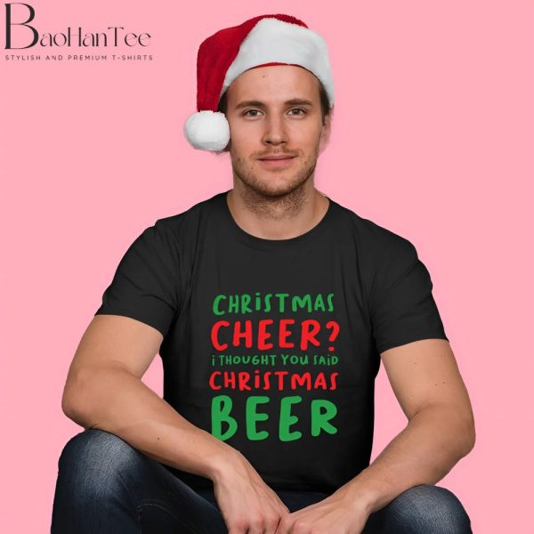 A man wearing a black T-shirt with a festive design that reads "Christmas Cheer? I thought you said Christmas Beer" in red and green text. He is also wearing a Santa hat and sitting against a pink background.